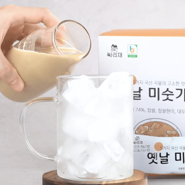 아침대용식으로 즐기기 좋은 옛날맛 미숫가루를 얼음 가득 담긴 투명 머그잔에 부어내는 장면. 곡물쉐이크 특유의 부드러운 질감이 유리잔 속으로 흘러 들어가며 시원하고 고소한 한 끼를 완성하는 모습.