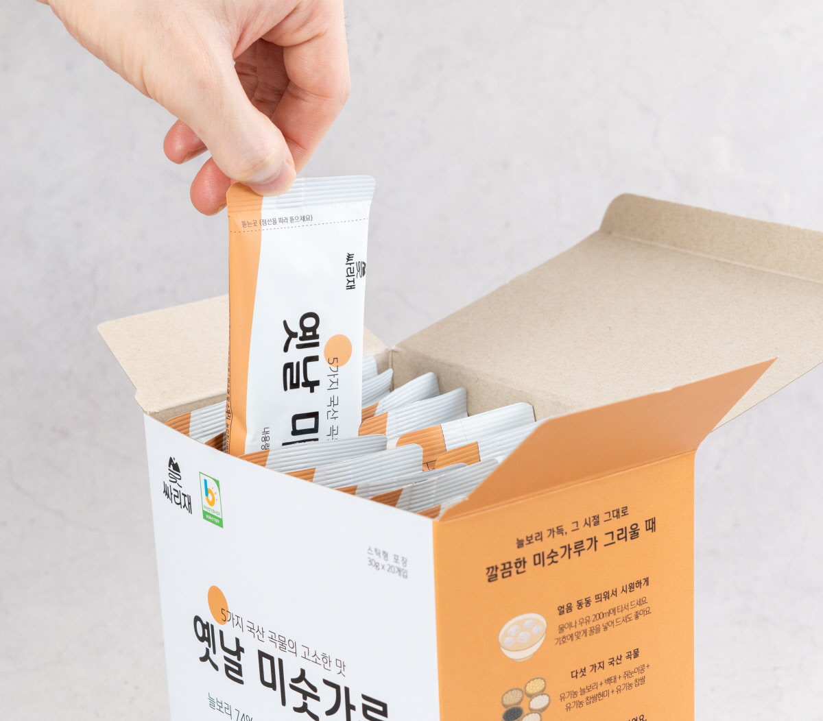 개별포장된 미숫가루 스틱을 한 손으로 꺼내는 모습. 깔끔한 디자인의 스틱포장이 여러 개 들어 있는 박스에서 간편하게 한 끼를 준비할 수 있는 식사대용선식의 활용성을 강조.