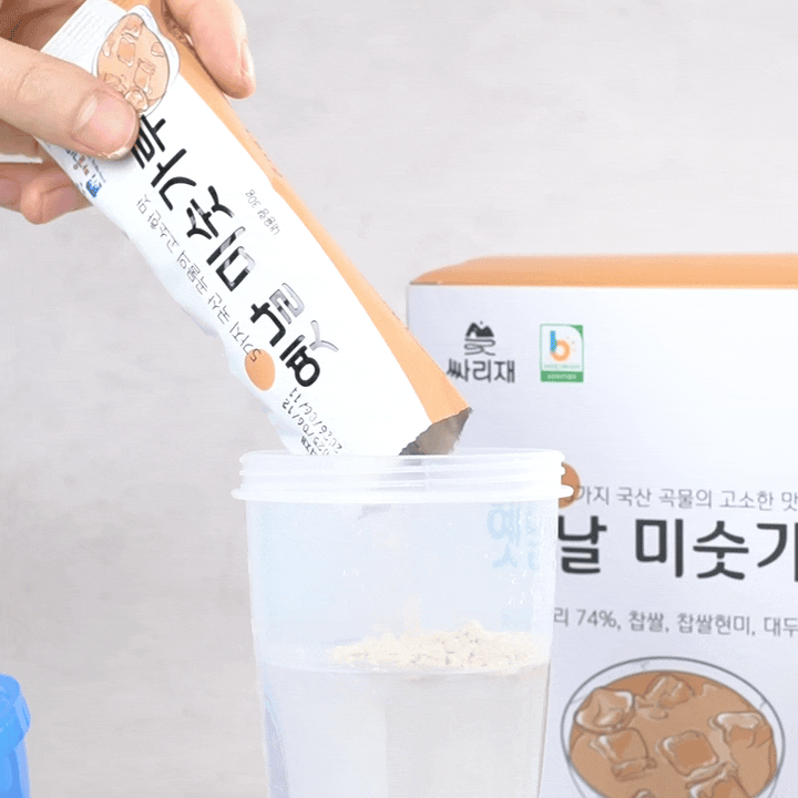 투명 컵에 미숫가루 스틱 한 포를 쏟아 붓는 모습. 물에 잘 풀리는 고운 곡물선식 가루가 담백한 아침대용식으로 간편하게 준비되는 과정을 보여줌.