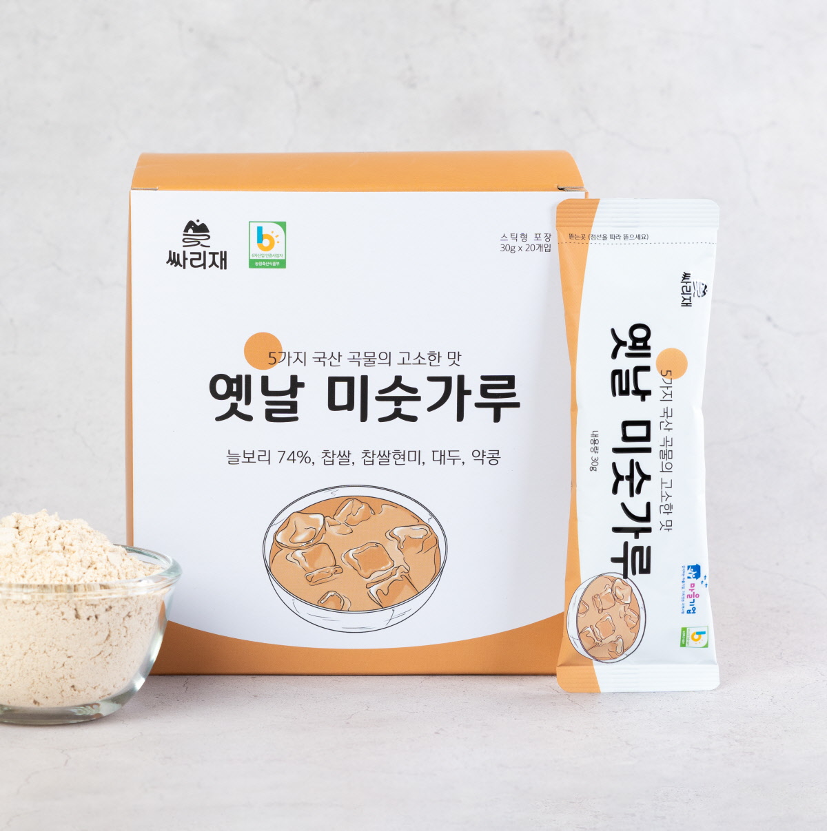 옛날 미숫가루 스틱과 제품 박스가 함께 놓여 있는 구성. 국산 약콩, 쥐눈이콩, 늘보리 등 5가지 국산 곡물로 만든 통곡물선식의 고소한 맛과 깔끔한 포장 디자인이 강조됨.