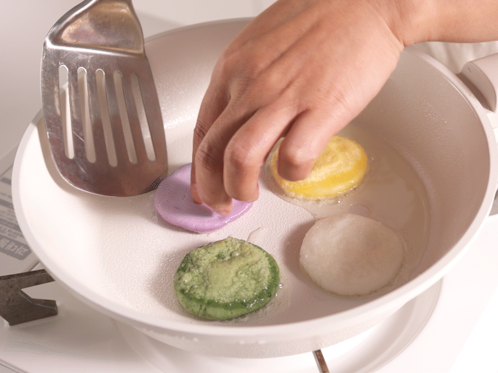 프라이팬 위에 다양한 색상의 화전이 놓이고 숟가락으로 기름을 두르며 굽는 장면으로, 메리골드꽃과 유기농현미반죽이 어우러진 화전만들기체험 과정을 보여준다.