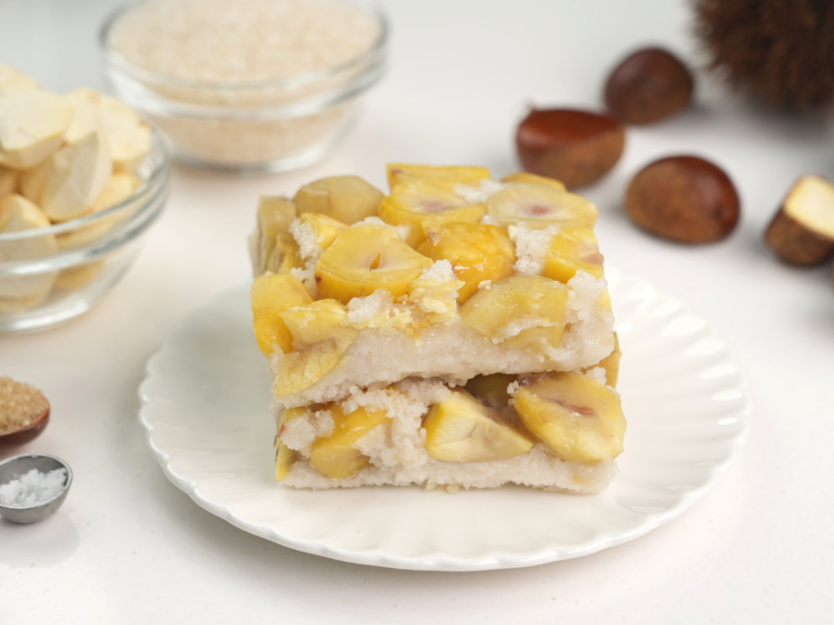 밤설기 건강간식 하얀 접시 위로 알밤이 듬뿍 박힌 두 장의 설기떡이 정갈하게 쌓여 있고 뒤로 유기농 설탕과 깐 밤이 흐릿하게 보이는 연출