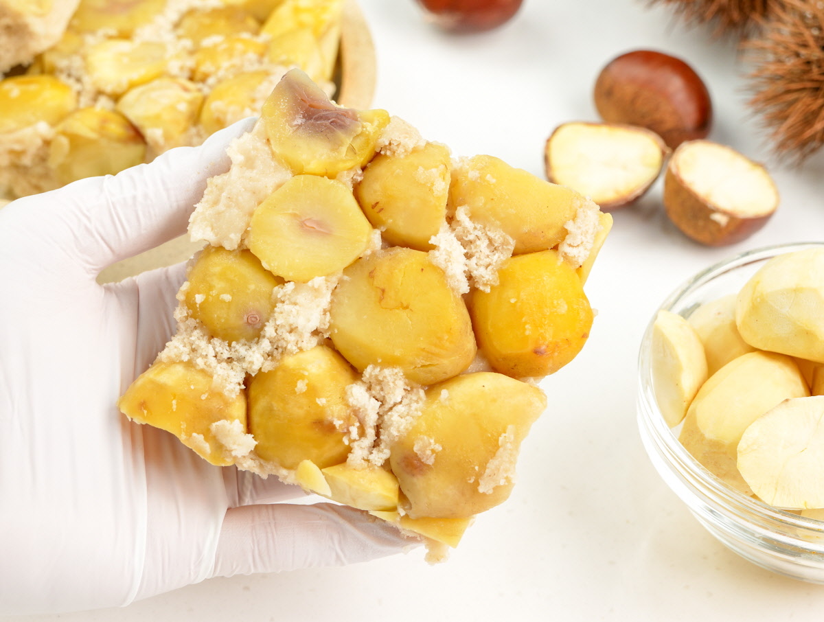 큼직한 국산 알밤이 듬뿍 박힌 현미 설기떡을 손으로 들어 올린 장면으로, 노란빛 밤 알갱이와 고운 떡결이 어우러져 담백한 건강간식의 질감을 느낄 수 있음