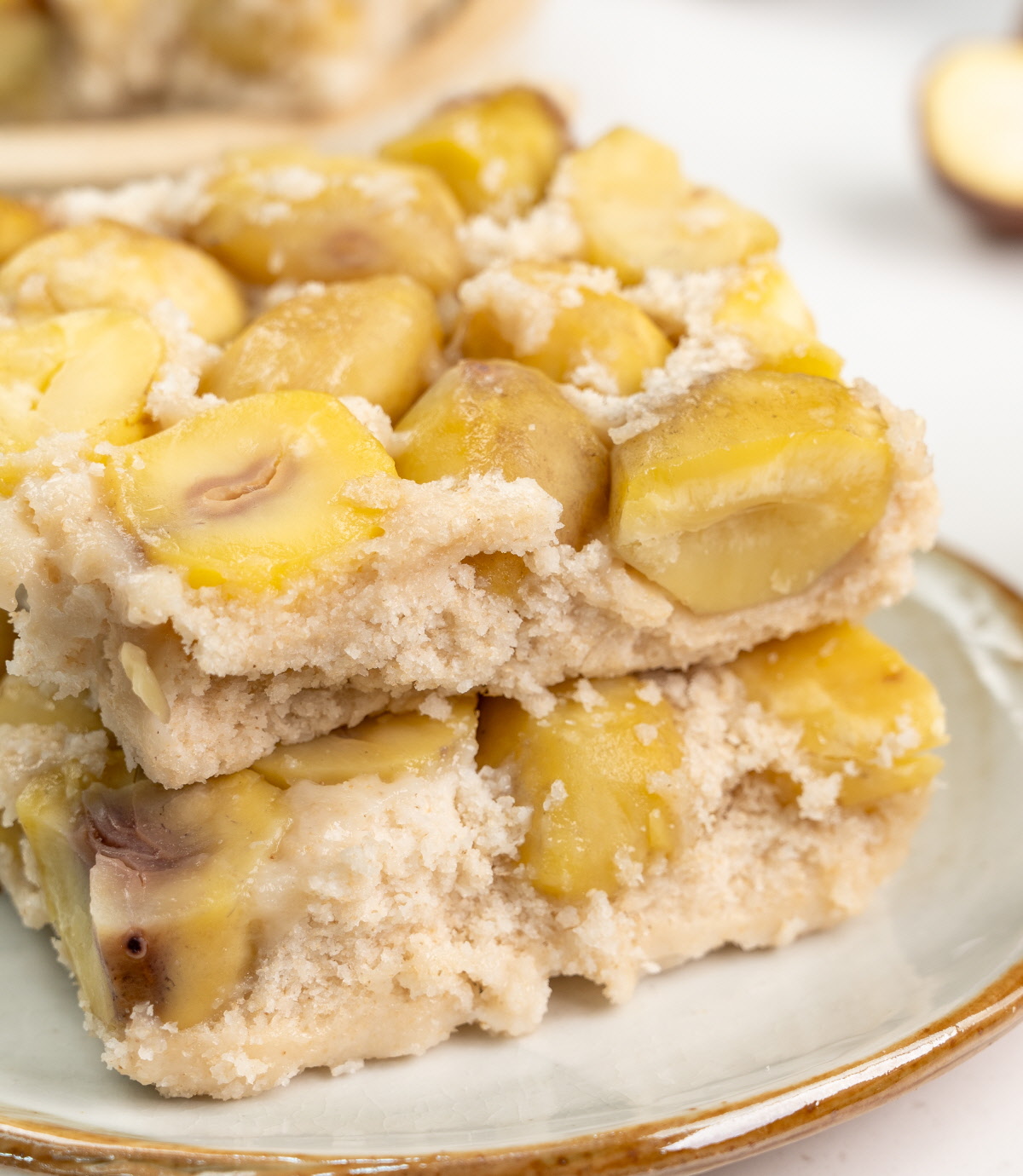 하얀 접시 위로 밤 알갱이가 듬뿍 박힌 현미 설기떡 두 조각이 정갈히 쌓여 있고 뒤편에는 깐 밤과 쌀이 어우러진 건강간식 연출