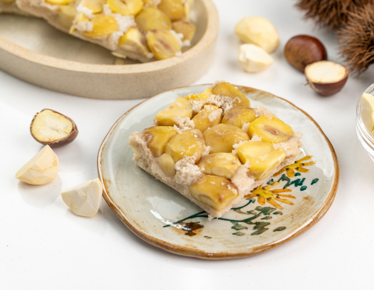 장갑 낀 손이 촉촉한 밤설기 조각을 들어 올려 알밤 알갱이와 부드러운 설기층이 근접하게 보이는 아침대용 간식 연출