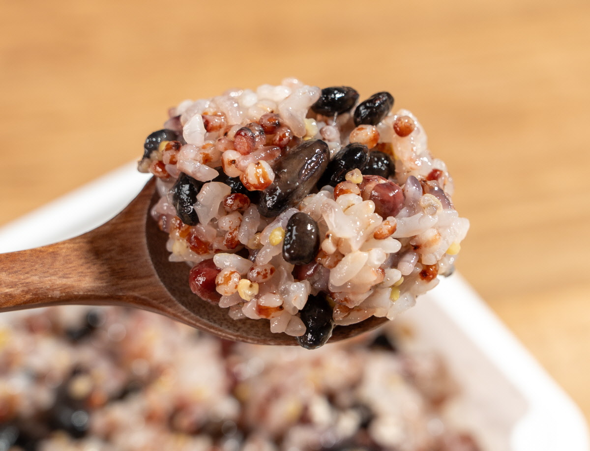 오곡찰밥 간편식 나무 숟가락 위에 윤기 도는 찹쌀밥과 검은콩 붉은팥 조잡곡 알갱이가 섞여 한입 크기로 떠진 모습, 뒤로 흰 플라스틱 용기 속 밥이 흐릿하게 보임