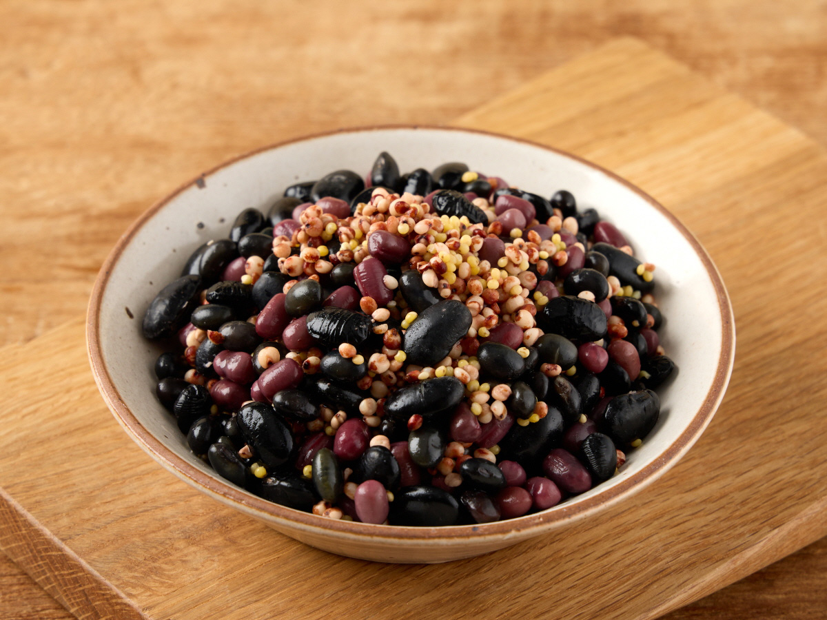 오곡찰밥 간편식 재료로 보이는 검은콩과 붉은팥, 기장 수수 같은 잡곡이 섞여 도자기 그릇에 담긴 모습, 나무 도마 위에 놓임