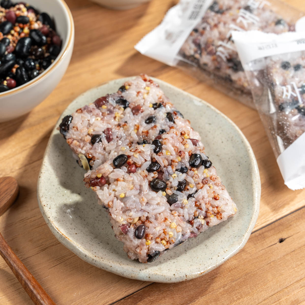 간편 영양식 오곡밥 담음새 흰 접시 위에 네모 모양 잡곡밥이 놓이고 옆에 싸리재 떡 문구가 보이는 개별포장이 함께 배치된 상단 구도