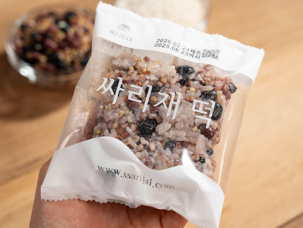 간편 영양식 오곡밥 개별포장 한 봉을 손에 들고 있으며 투명 포장 안에 검은콩 붉은팥 잡곡이 섞인 찹쌀밥이 보인다