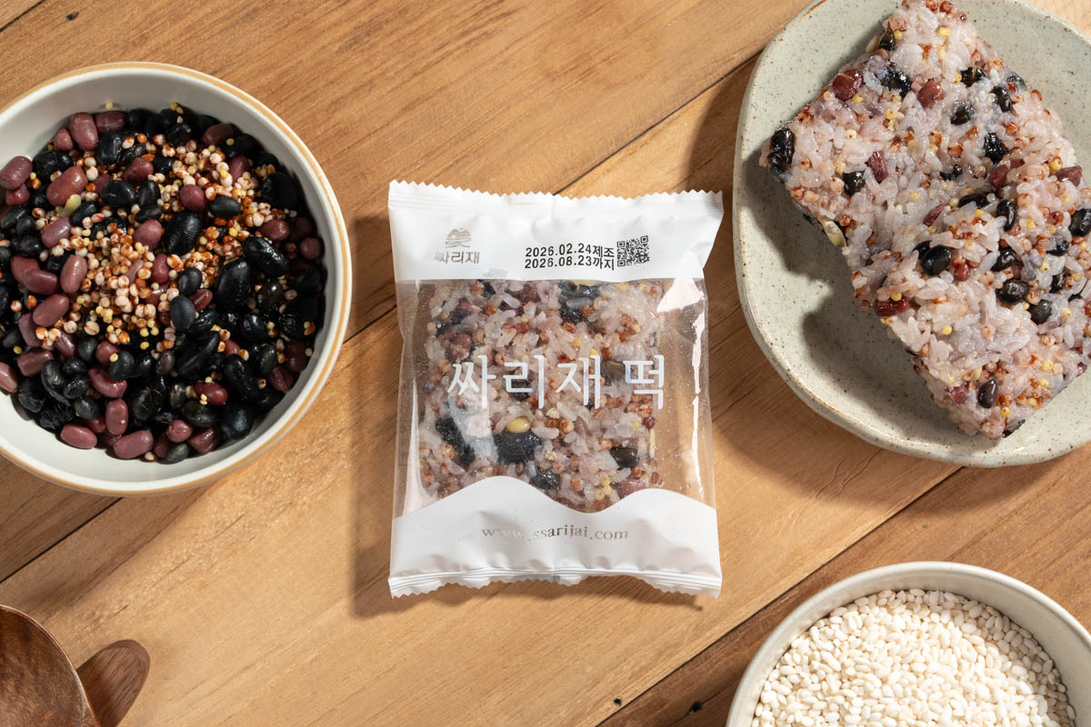 간편 영양식 오곡밥 구성 컷 나무 테이블 위에 콩과 잡곡이 담긴 그릇, 흰쌀이 담긴 그릇, 중앙에 싸리재 떡 문구가 보이는 개별포장, 오른쪽 접시에 네모 모양으로 빚은 잡곡밥이 함께 놓임