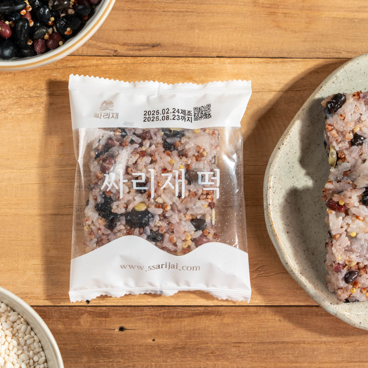 간편 영양식 오곡밥 개별포장 한 봉이 나무 테이블 위에 놓여 있고 투명 포장 안에 잡곡밥과 콩이 보이며 전면에 싸리재 떡, 하단에 www.ssarijai.com 표기가 선명함