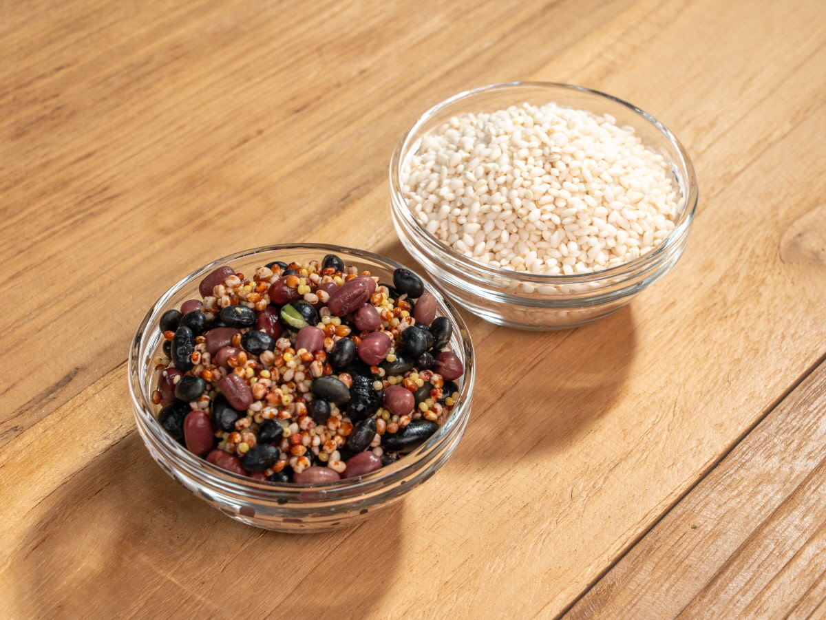간편 영양식 오곡밥 재료 연출로 보이는 유리 그릇 두 개에 검은콩 붉은팥 잡곡 혼합물과 흰쌀이 각각 담겨 나무 테이블 위에 놓인 모습