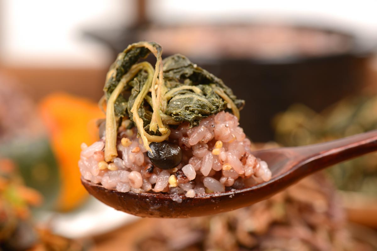 오곡찰밥 간편식 나무 숟가락 위에 잡곡밥 한입과 검은콩이 보이고, 위에 데친 나물 줄기와 잎이 올려져 있다