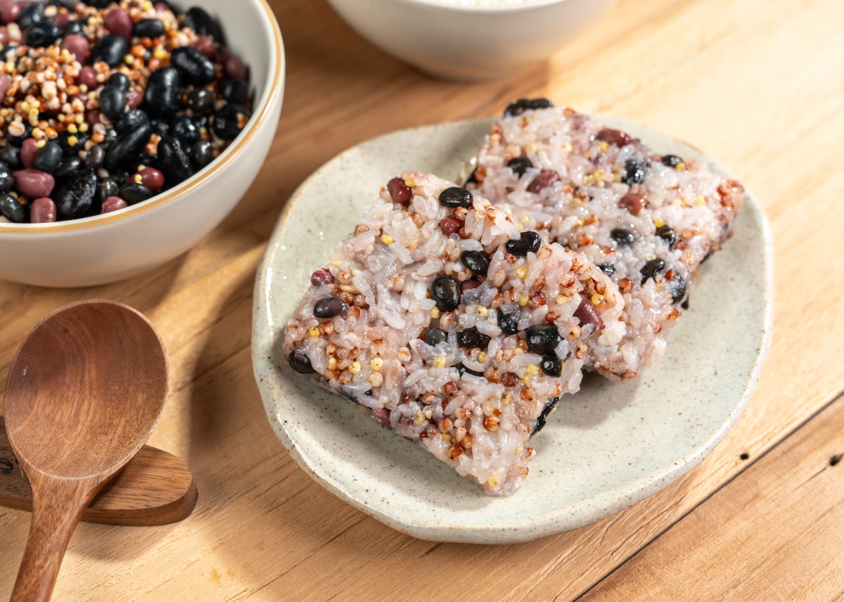 간편 영양식 오곡밥 네모 모양으로 빚은 잡곡밥 두 조각이 회색 접시에 겹쳐 담겨 있고, 밥알 사이로 검은콩 붉은팥 노란 잡곡이 고르게 섞여 보이며 옆에 나무 숟가락과 뒤쪽에 콩잡곡 그릇과 흰쌀 그릇이 함께 놓임