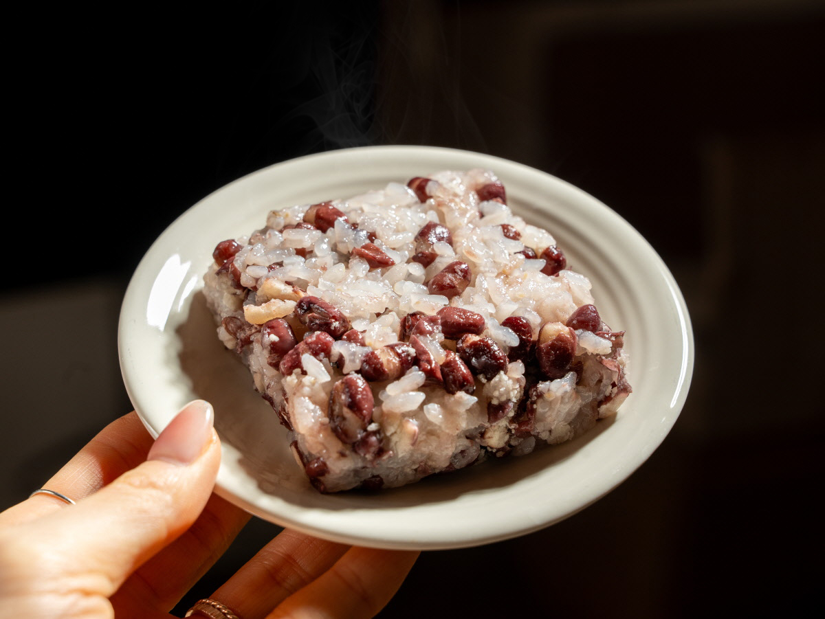 따끈하게 데워져 김이 모락모락 나는 팥 찰밥을 접시에 담아 손으로 들고 있는 모습