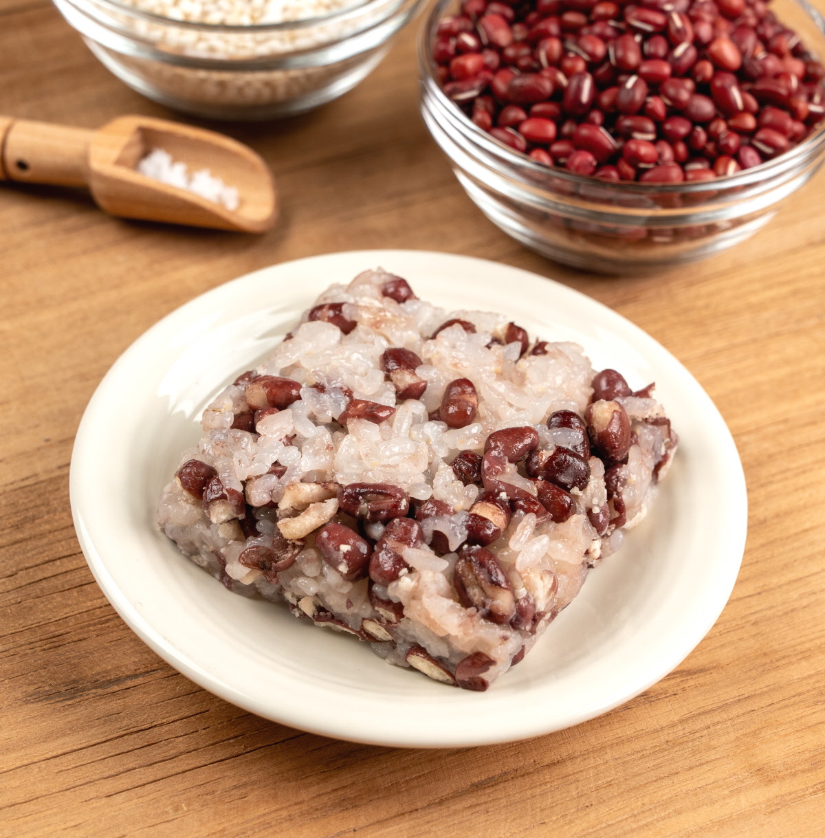 밝은 배경의 나무 테이블 위 접시에 담긴 팥 찰밥과 잡곡 재료들의 조화로운 전체 컷