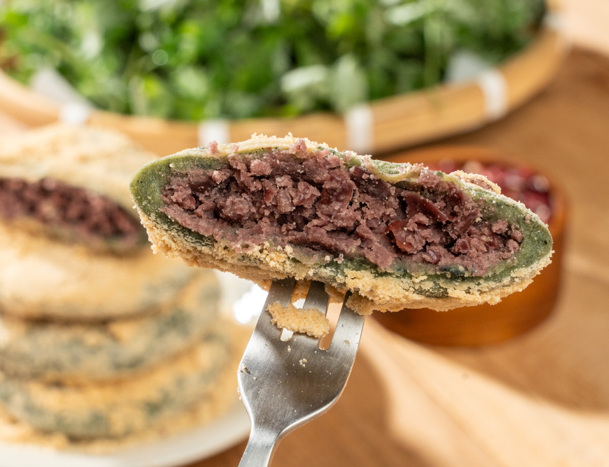 앙금쑥개떡 싸리재 개떡 한 조각을 포크에 올려 가까이 보여주는 장면으로 얇은 쑥떡 피 안에 통팥앙금이 가득 차 있고 표면에는 콩고물이 고루 묻어 있는 모습