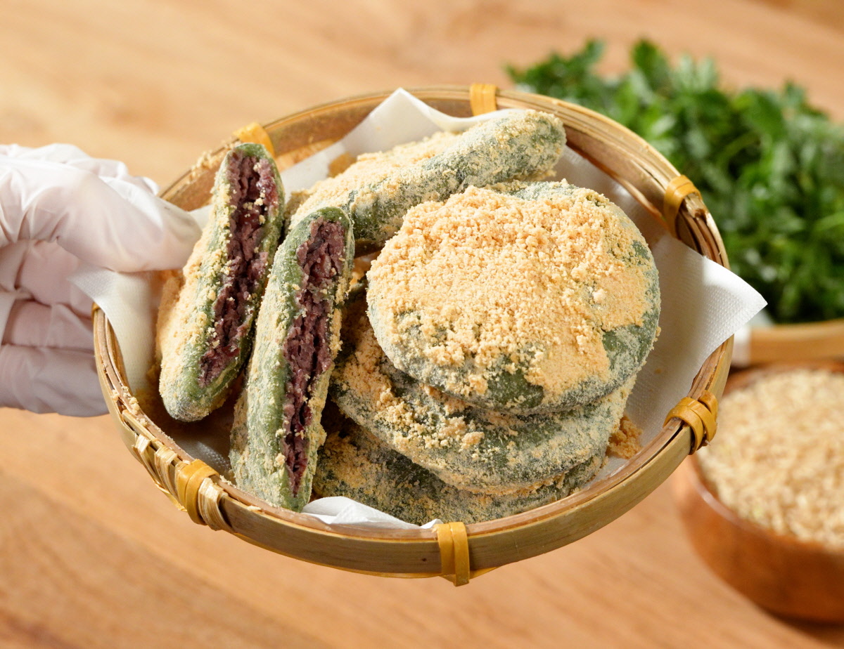 앙금쑥개떡 싸리재 개떡을 작은 채반에 담아 손으로 들어 올린 모습으로 통팥앙금 단면 두 조각과 콩고물 묻은 둥근 떡 여러 개가 함께 담긴 구성