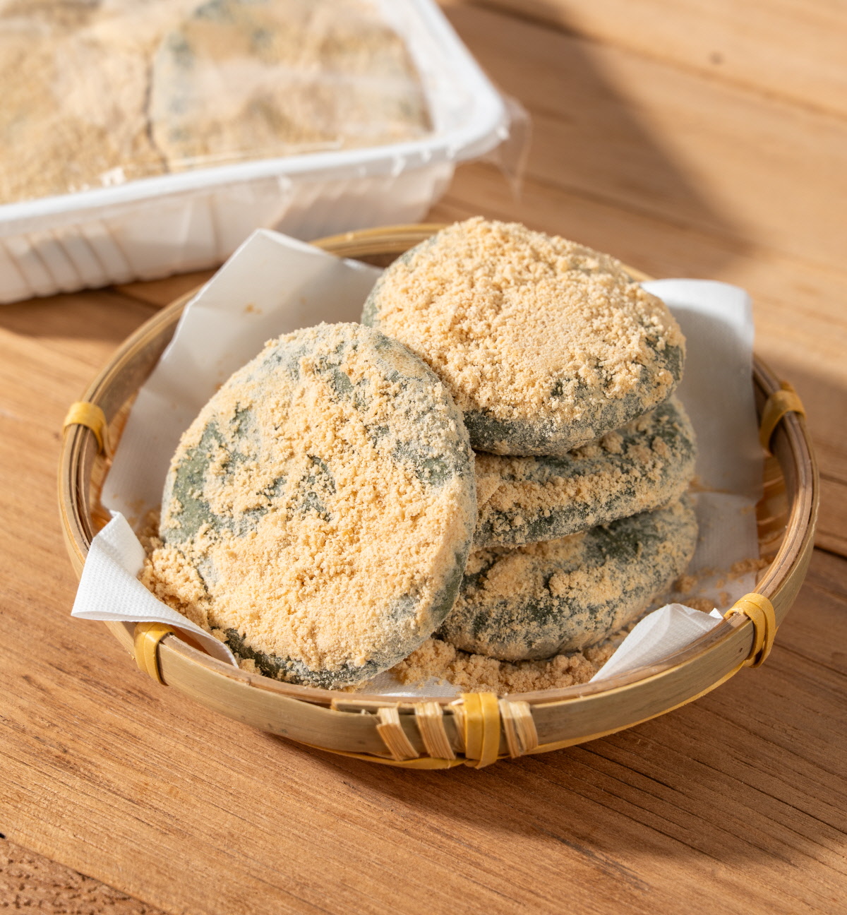 앙금쑥개떡 싸리재 개떡 여러 개를 둥근 채반에 담아 놓은 모습으로 겉면에 콩고물이 넉넉히 덮여 있고 뒤쪽에는 포장 용기에 담긴 제품이 함께 보이는 구성