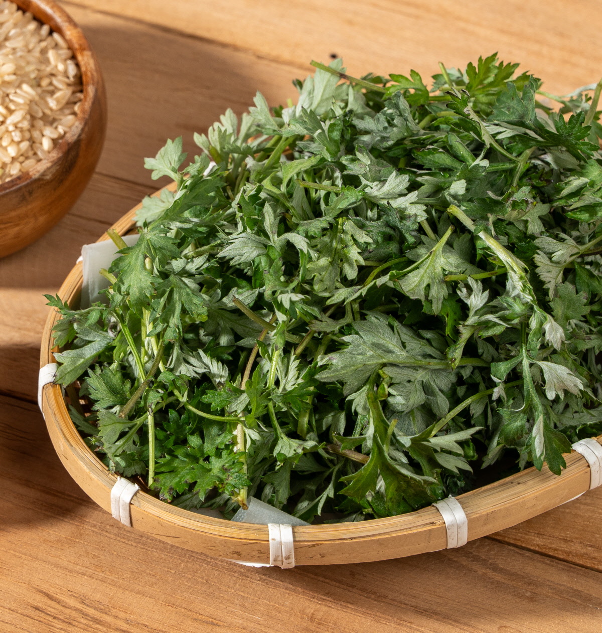 쑥개떡 싸리재 개떡에 사용하는 어린 쑥을 대나무 채반에 수북하게 담아 놓은 모습으로 짙은 초록빛 잎과 부드러운 줄기가 가득 보이고 옆에는 현미가 담긴 그릇이 일부 함께 놓여 있는 구성