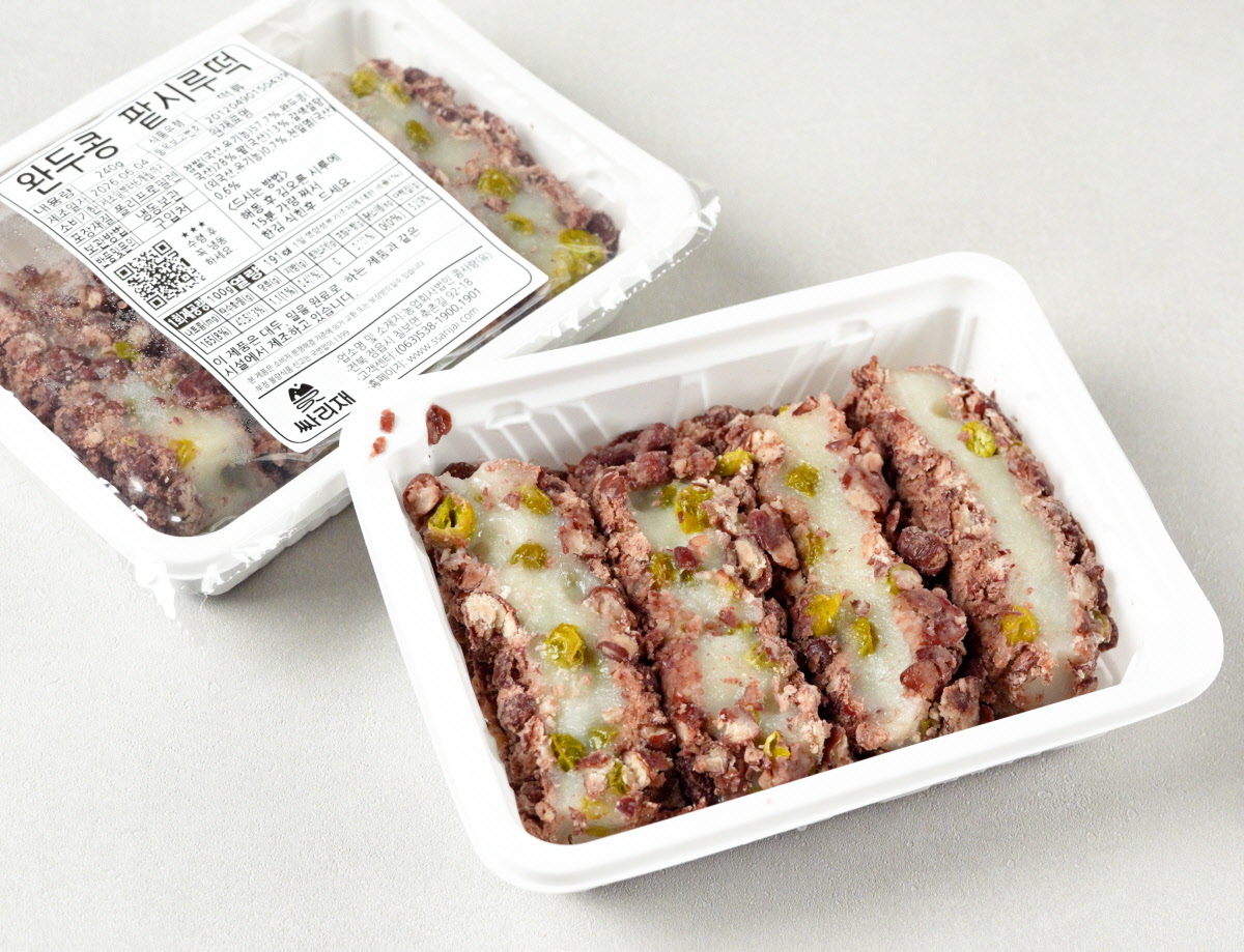 완두콩 고물이 들어간 팥시루떡 두 팩이 깔끔한 회색 바탕 위에 놓여 있습니다. 앞쪽 팩은 밀봉이 해제되어 큼직하게 썰린 떡 네 조각의 단면을 그대로 보여주며 뒤쪽 팩은 완두콩 팥시루떡이 깔끔하게 패키징되어 있습니다.