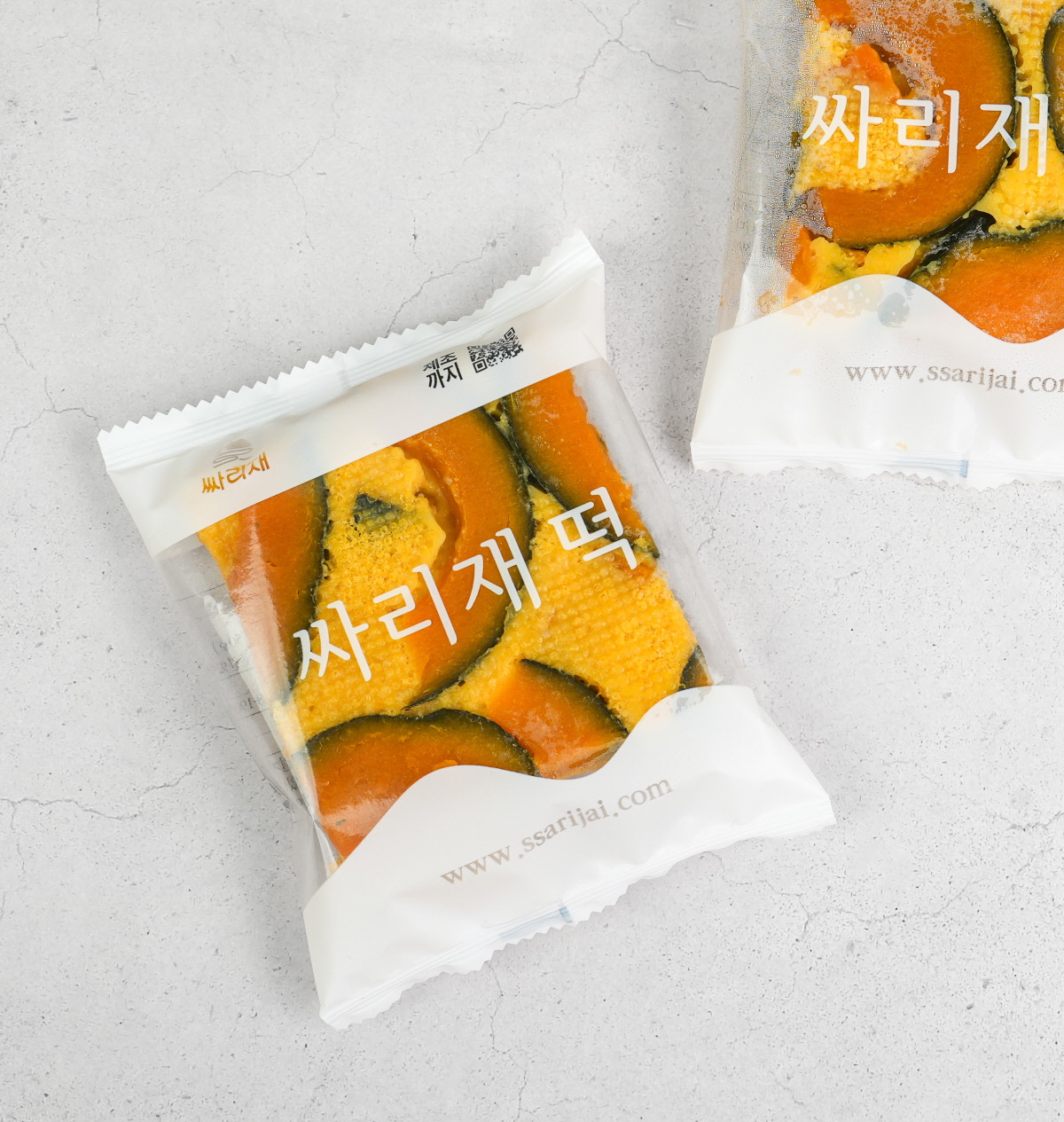 싸리재 단호박 설기 떡이 접시에 담겨 있고, 옆에는 브랜드 로고가 박힌 개별 포장 떡 3개가 나란히 놓여 있다. 낱개 포장으로 위생적으로 보관할 수 있는 냉동떡 형태로, 간편한 아침 간식으로 활용하기 좋다.