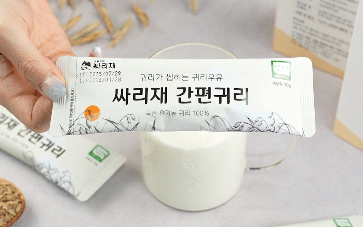 간편귀리 스틱 포장을 손에 들고 흰 우유가 담긴 컵 위로 보여주는 장면으로, 국산 유기농귀리 100% 제품이 깔끔하게 디자인된 소포장 형태