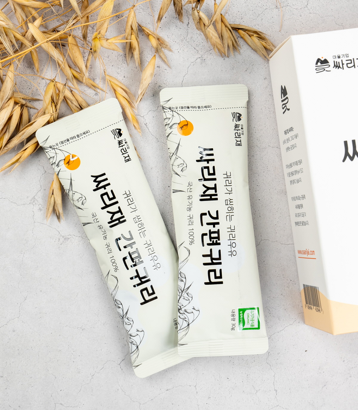 회색 배경 위에 놓인 유기농귀리 간편 스틱 2개와 납작귀리 알곡, 아침대용 귀리모닝으로 활용 가능한 국산귀리 제품