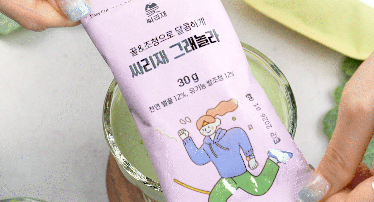 그래놀라 스틱 포장을 손으로 잡아 들고 유리잔 위에 올린 모습, 연보라색 패키지에 ‘꿀&조청으로 달콤하게 30g, 천연 벌꿀 12%, 유기농 쌀조청 12%’ 문구가 인쇄되어 있으며 귀리와 통곡식의 건강한 간식을 강조