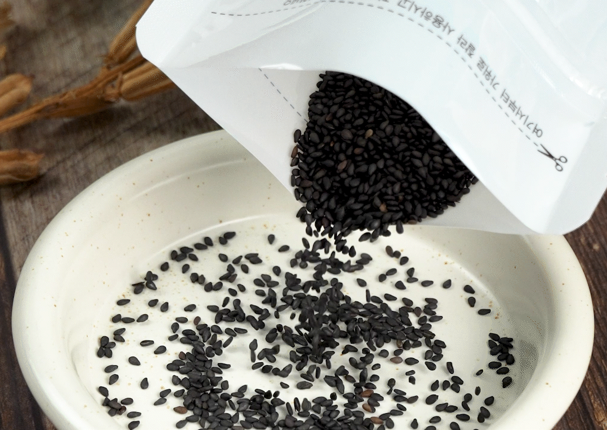 흑임자와 유기농깨 향이 살아 있는 볶은깨가 하얀 그릇 위로 부드럽게 떨어지며 퍼지는 모습. 고운 검정깨 알갱이가 고소한흑임자 특유의 윤기를 띠고 흩어져 있는 장면.