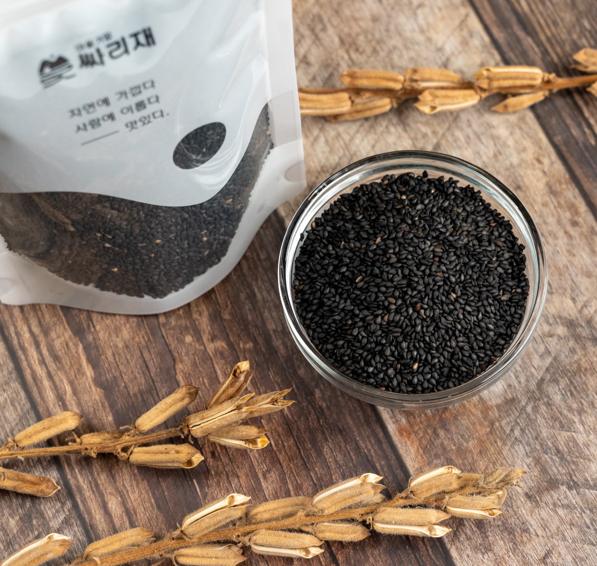 국산흑임자와 유기농깨로 만든 볶은깨가 투명한 볼에 가득 담겨 있으며, 옆에는 싸리재 포장이 서 있고 주변에 검정깨 꼬투리가 놓여 자연스러운 분위기를 주는 장면.