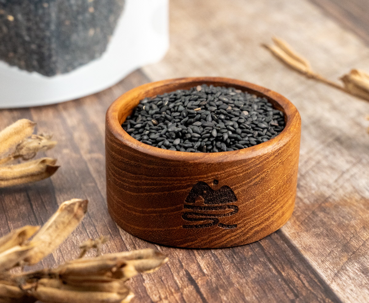 잘 볶은 국산흑임자가 작은 나무 그릇에 가득 담겨 있으며, 주변에 말린 깨 꼬투리가 놓여 고소한 흑임자가루 원료의 자연스러움을 강조한 장면.