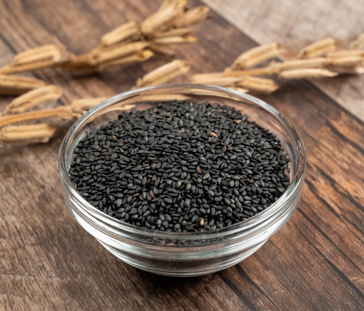 흑임자와 국산검정깨가 투명한 유리볼에 듬뿍 담겨 있으며, 뒤쪽에는 마른 깨 꼬투리가 놓여 고소한흑임자 원물의 질감을 강조한 구성.