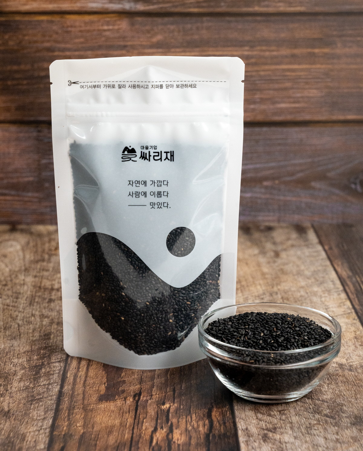 유기농깨로 만든 흑임자 원물이 담긴 포장팩이 세워져 있고, 앞에는 국산검정깨가 유리볼에 담겨 놓인 모습으로 자연스러운 포장 비주얼을 보여주는 장면. 