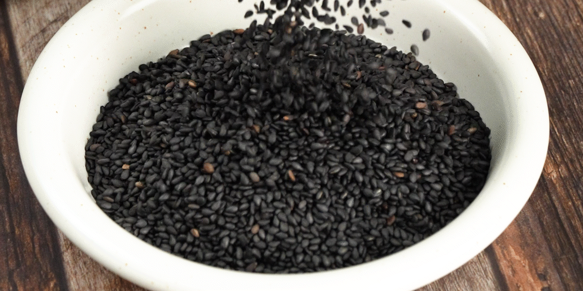 국산깨로 볶아낸 검정깨가 하얀 그릇 속으로 부드럽게 쏟아지며 담기는 장면. 고소한흑임자의 윤기와 볶은깨 특유의 알갱이감이 생생하게 표현됨.