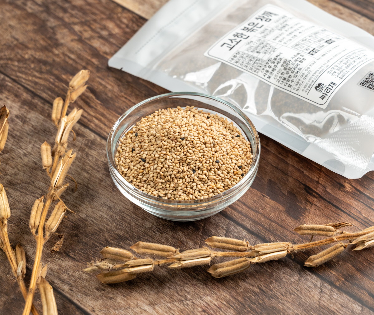 투명 유리볼에 국내산통깨가 가득 담겨 있고 주변에 참깨 꼬투리가 놓여 있어 유기농참깨의 자연스러운 원물 느낌을 보여주는 구성.