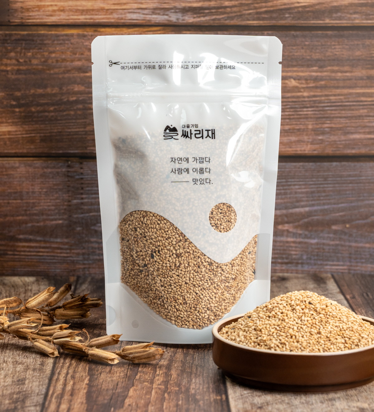 참깨가 듬뿍 담긴 대용량 포장팩이 세워져 있으며 앞에는 볶은참깨가 담긴 그릇이 놓여 원물의 색감과 고소함을 강조한 장면. 