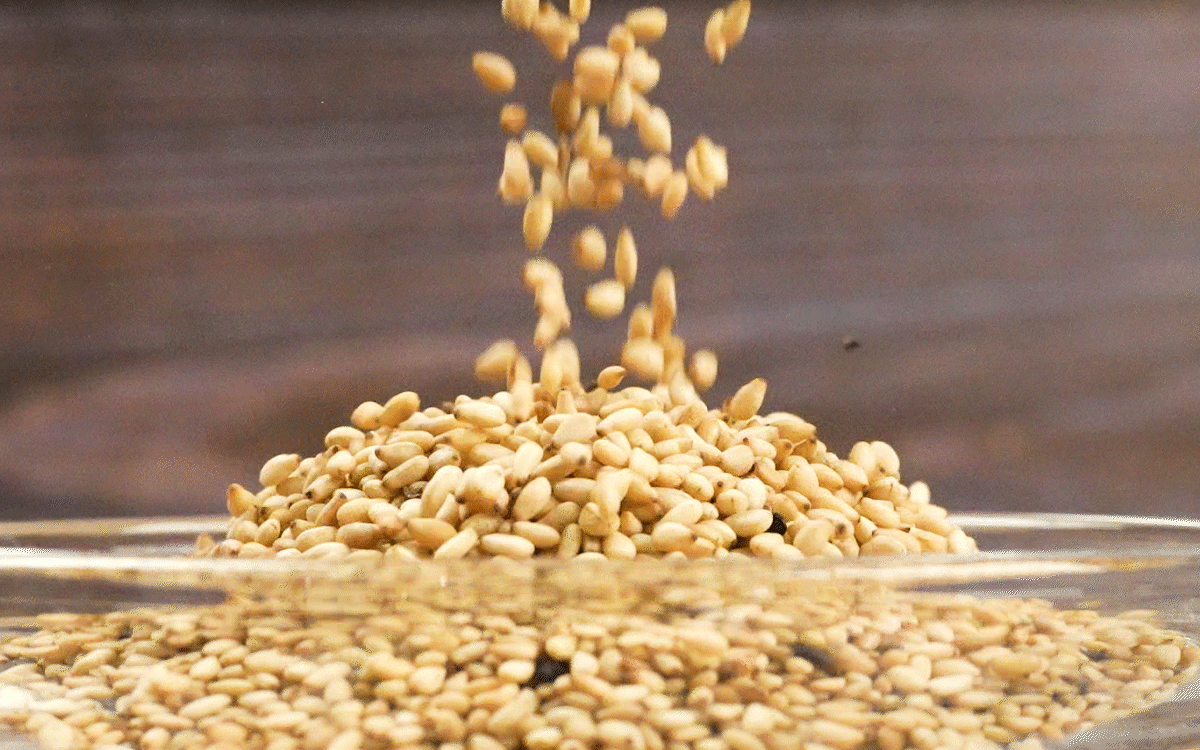 볶은참깨와 고소한참깨가 위에서 떨어져 투명한 그릇 안에 둥글게 쌓이며, 국산참깨 알갱이가 반짝이는 질감을 보여주는 장면.