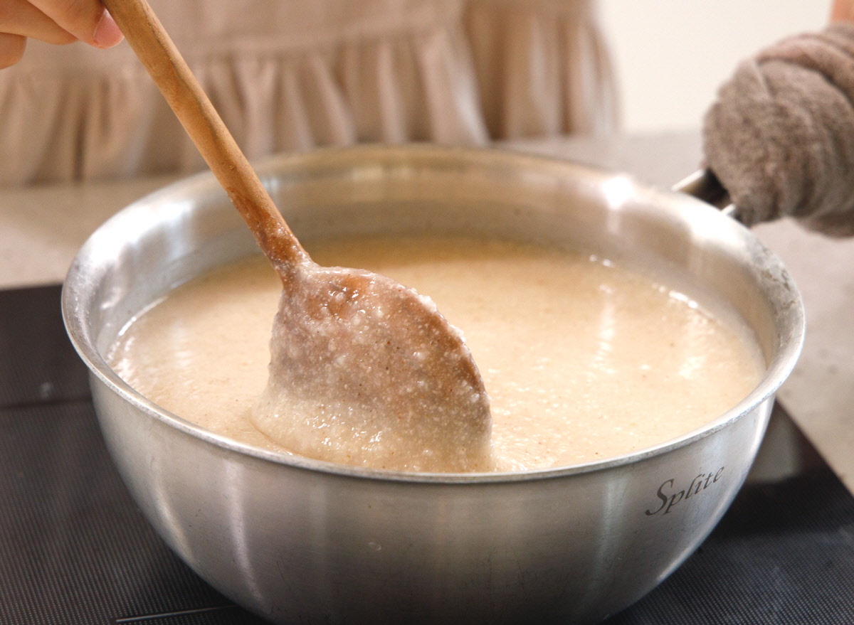 따뜻하게 끓인 참깨죽을 스테인리스 냄비에서 나무주걱으로 저어 올리는 모습으로, 볶은참깨와 국산참깨의 고소함이 잘 우러난 부드러운 죽이 걸쭉하게 담겨 있음.