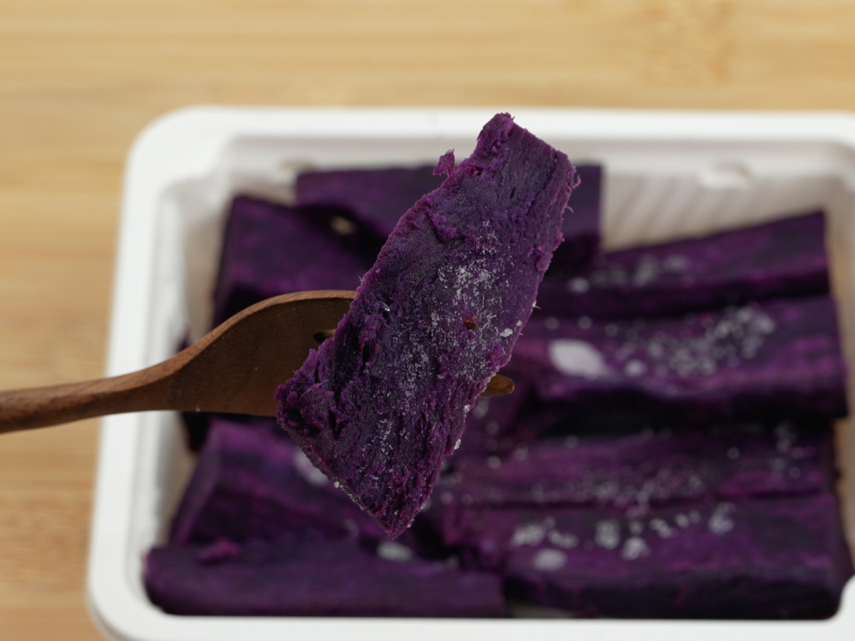 나무 포크에 꽂은 자색고구마 조각을 클로즈업한 장면으로, 표면에 하얀 결정이 맺힌 듯한 질감이 보이며 구운 고구마의 단단한 결이 표현됨