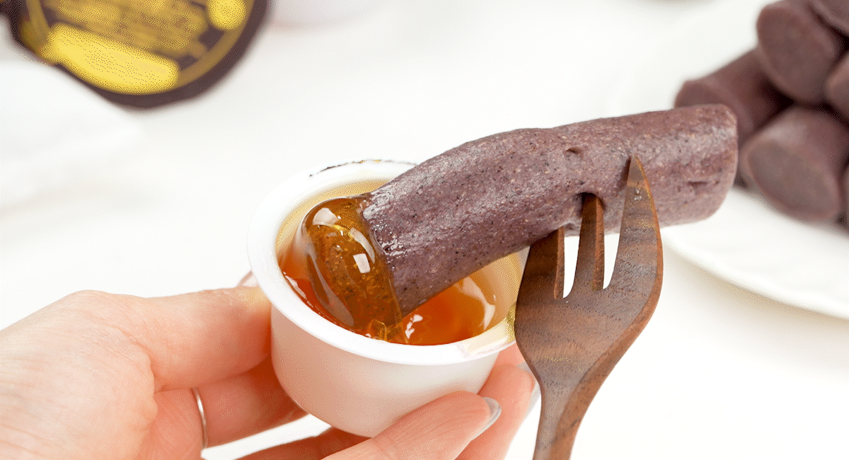 쫀득한 흑미 가래떡을 포크로 들어 황금빛 꿀에 찍은 장면으로, 고소한 현미떡의 질감과 흑미의 은은한 색감이 어우러져 담백한 건강간식으로 느껴진다.