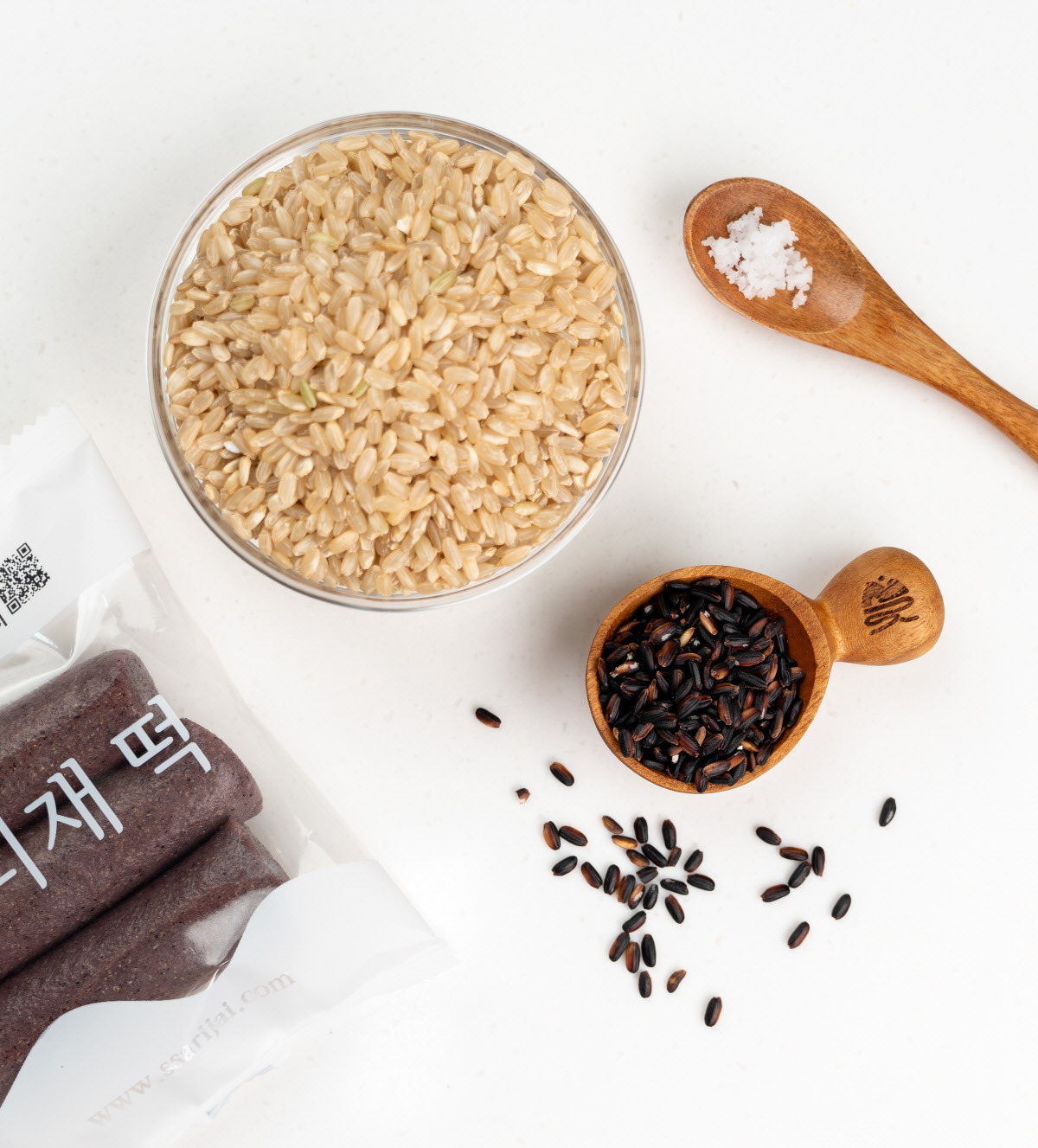 투명한 개별포장 속 흑미 가래떡이 정갈하게 담긴 모습으로, 뒷배경에는 흑미와 현미, 천일염 재료가 함께 놓여 자연스러운 건강간식 이미지를 완성했다.