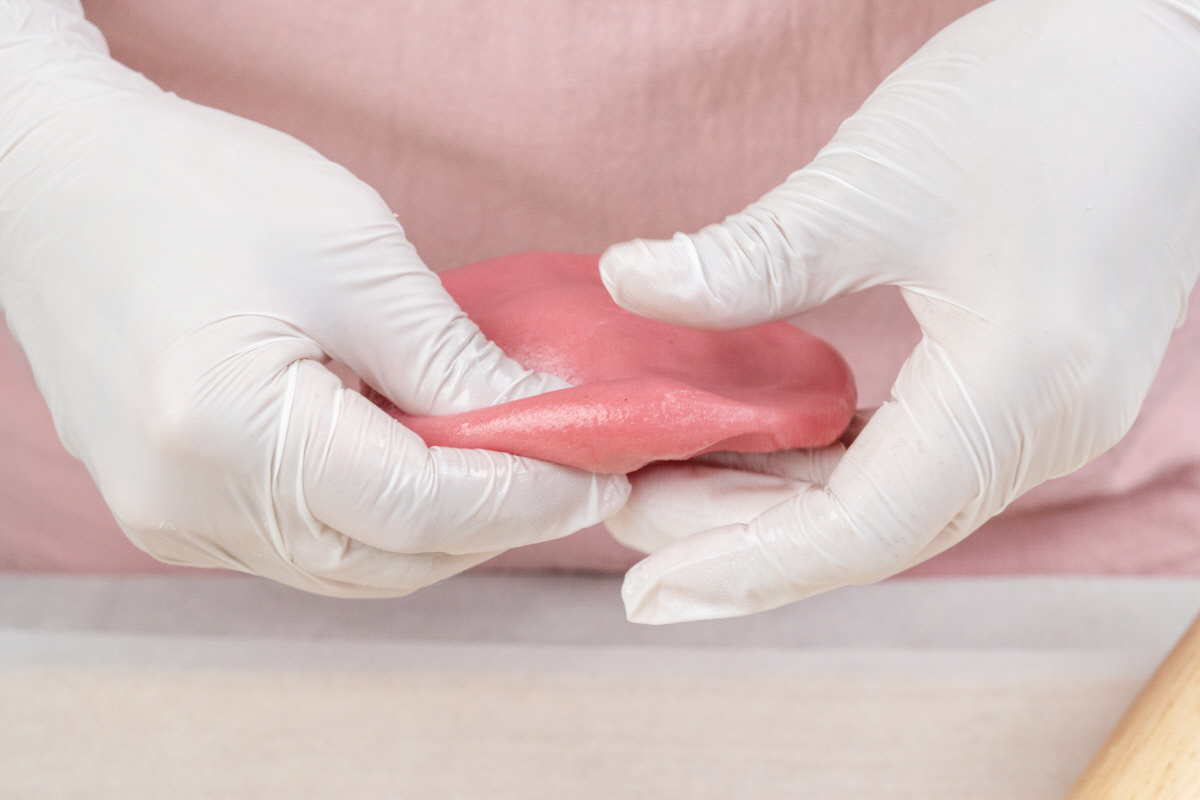 위생장갑을 낀 손으로 분홍색 떡 반죽을 펼쳐 쥐고 있는 모습으로, 말랑하고 부드러운 질감이 강조되며 카네이션바람떡 만들기 준비 단계의 섬세한 작업 과정을 보여준다.