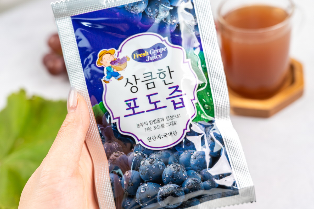 손에 든 상큼한 포도즙 파우치 손에 들린 파우치 정면에 ‘상큼한 포도즙’, ‘농부의 땀방울과 정성으로 키운 포도를 그대로’, ‘원산지: 국내산’ 문구가 보이고 뒤에는 유기농포도즙 한 잔이 놓여 있다.