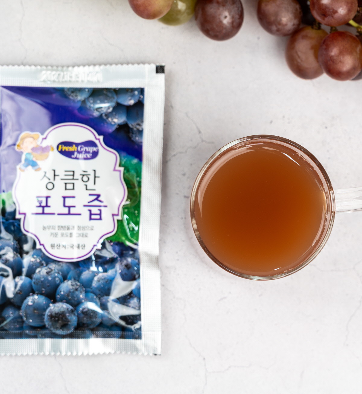 파우치와 잔을 나란히 둔 포도즙 ‘상큼한 포도즙’ 파우치와 함께 투명한 잔에 갈색빛 유기농포도즙이 나란히 놓여 있고, 위쪽에는 유기농포도 송이가 보여 한 봉을 따라 마시는 구성을 표현한다.
