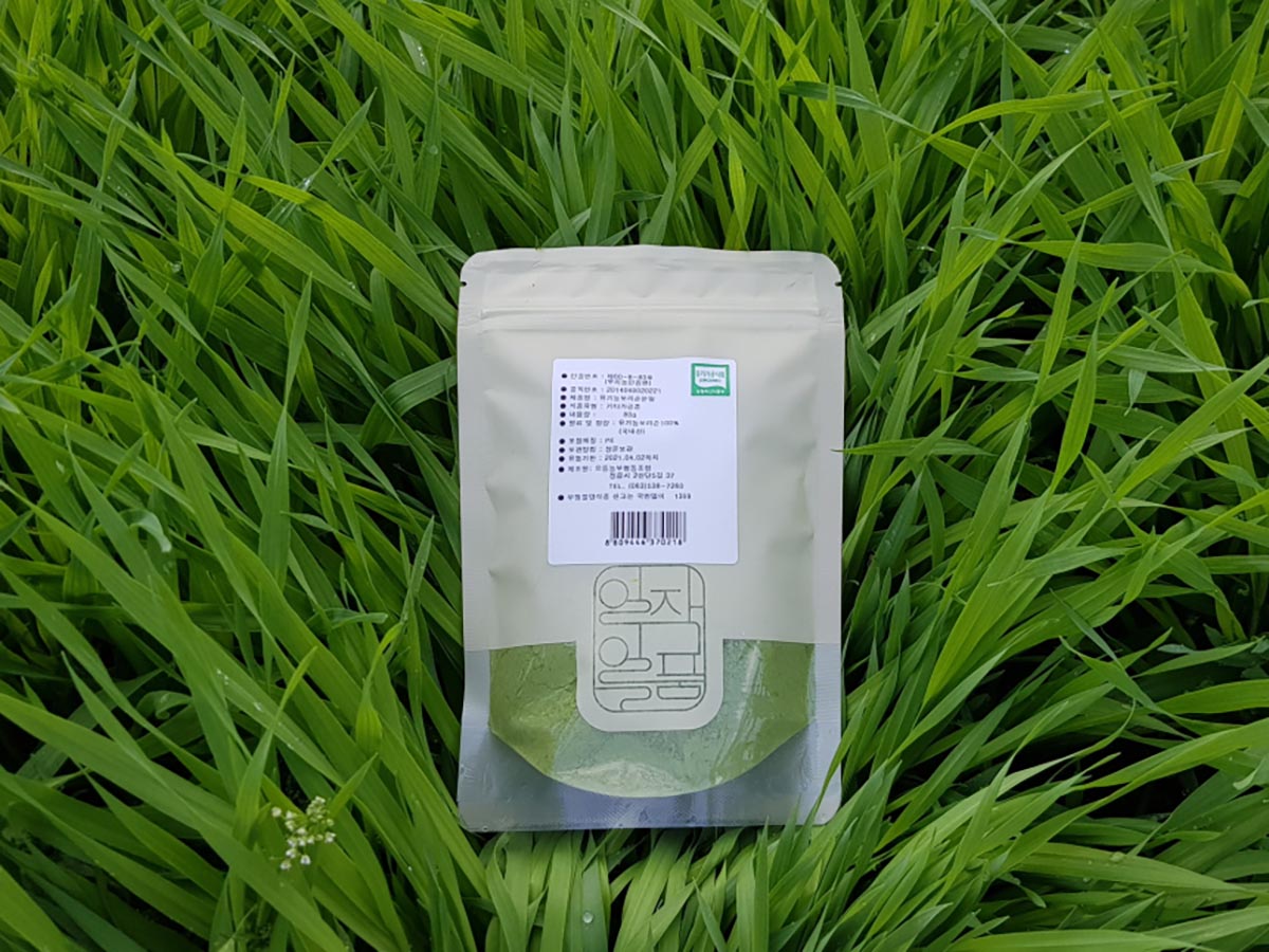초록빛 보리밭 위에 비닐 포장지에 포장된 보리순 분말 제품이 눕혀져 있습니다.