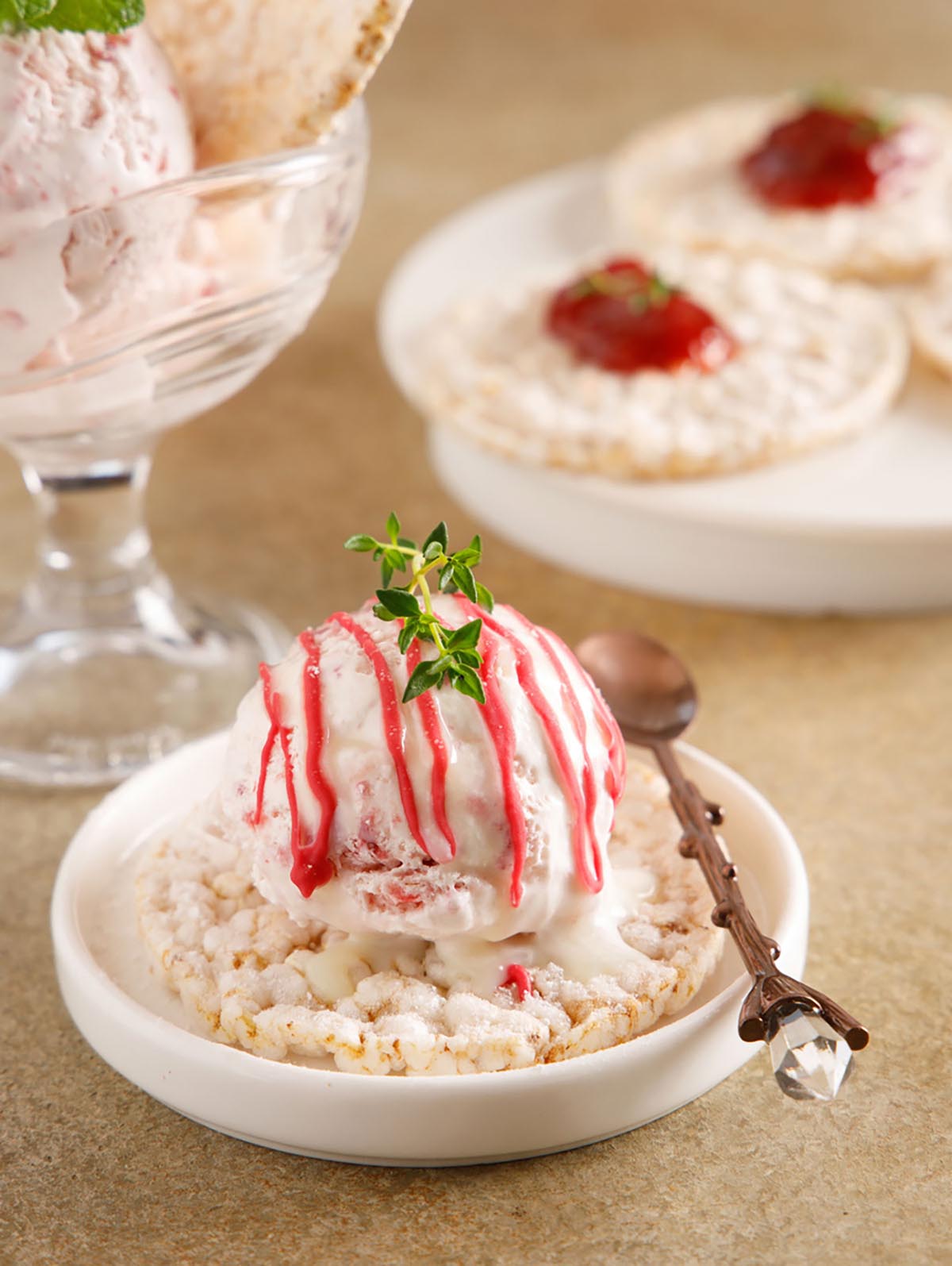  하얀 접시에 현미과자 한개가 있고, 그 위에 아이스크림 한스푼이 올려져 있다.아이스크림 위에 새싹잎으로 장식을 더하고 수저가 있다.