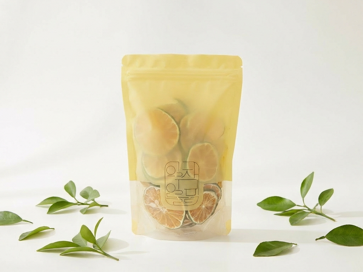 과일칩 천연간식이 담긴 노란 스탠드 지퍼 파우치가 흰 배경 중앙에 세워져 있고 아래쪽 투명 부분으로 동그란 과일칩이 비치며 주변 바닥에 초록 잎 장식이 흩어져 있는 단독 연출