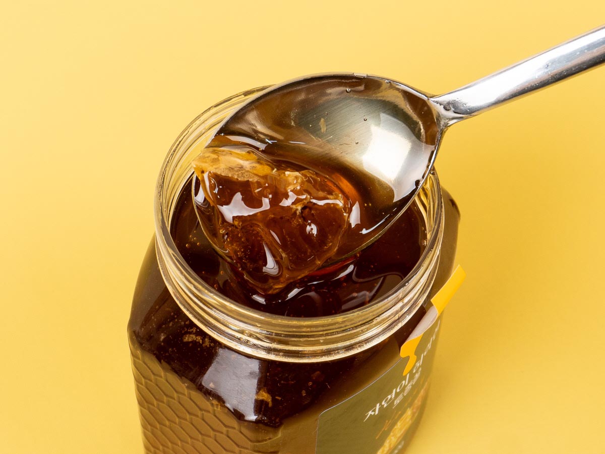 갈색의 토종벌꿀병을 열어 스텐숟가락으로 벌집조각과 꿀을 한수저 떠서 들어올리고 있습니다.