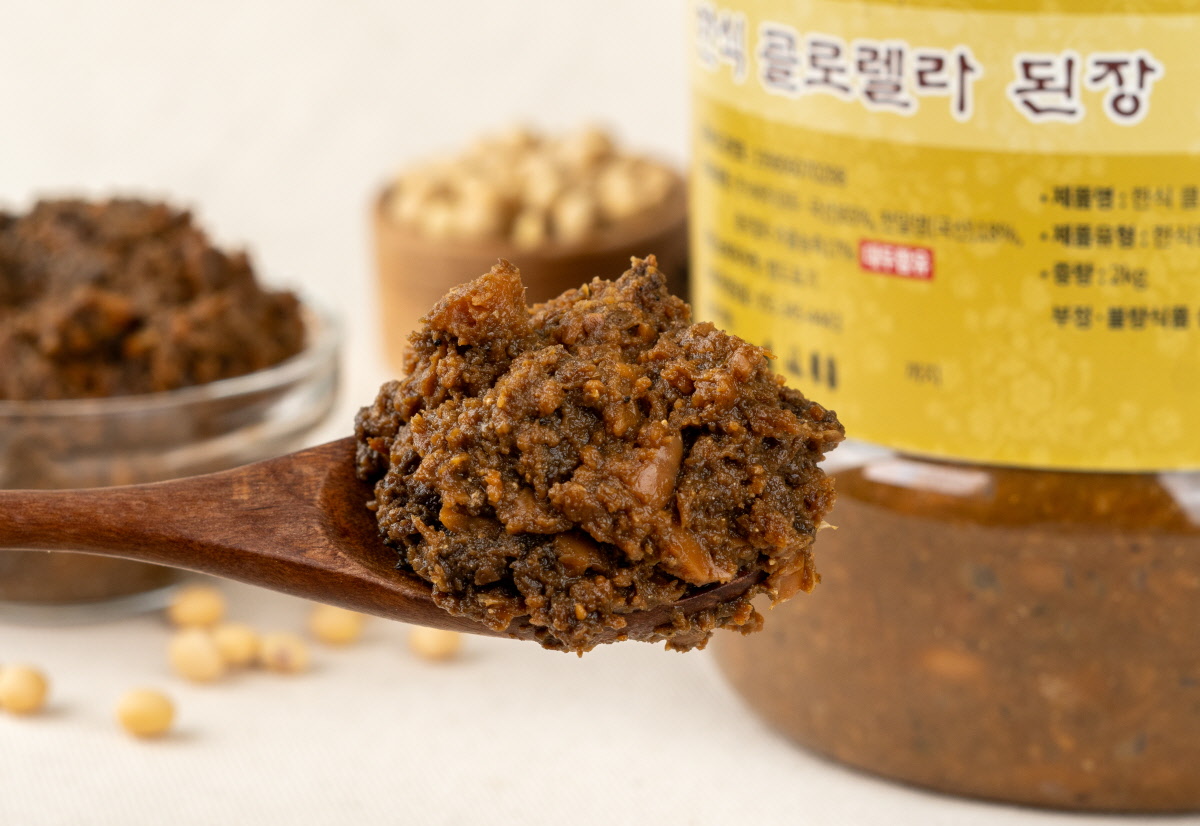 나무 숟가락에 퍼올린 클로렐라 집된장이 크고 굵은 메주 알갱이와 함께 담겨 있다. 뒷배경에는 된장 원통 용기와 국산 콩이 함께 보인다. 된장찌개용, 쌈장대신 두루 활용하기 좋다.